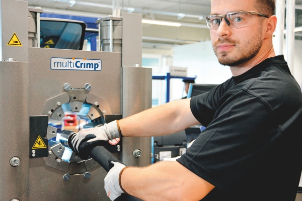 Mann mit Schutzbrille und Handschuhen bedient eine Hydraulikschlauchpresse der Marke "multiCrimp". Er hält einen Schlauch in die Maschine. Im Hintergrund ist eine Werkstattumgebung mit unscharfen Details erkennbar.  